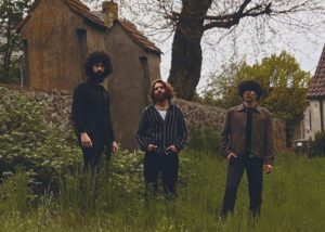 Three men in vintage clothing stand in a grassy field near old buildings.