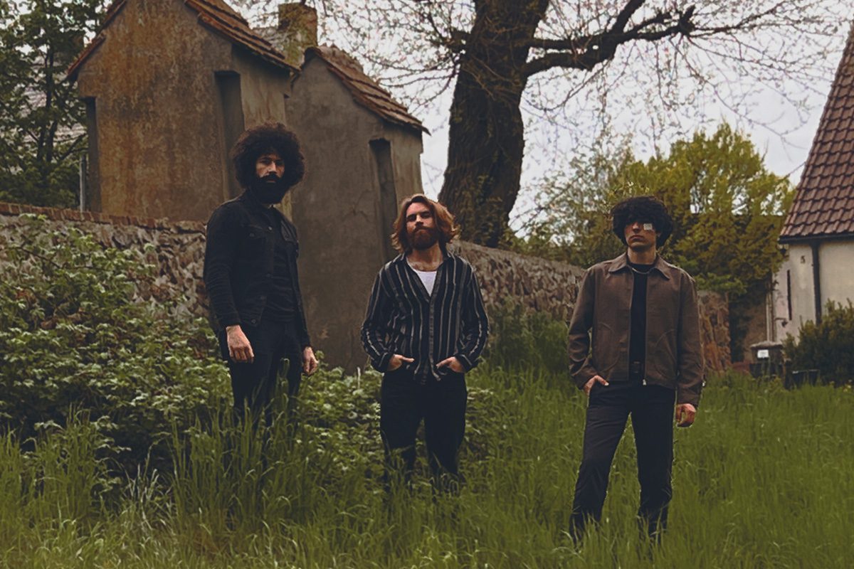 Three men in vintage clothing stand in a grassy field near old buildings.