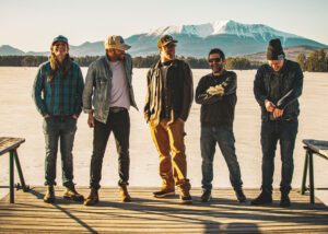 The Mallett Brothers band posing on a dock with a snowy mountain backdrop.